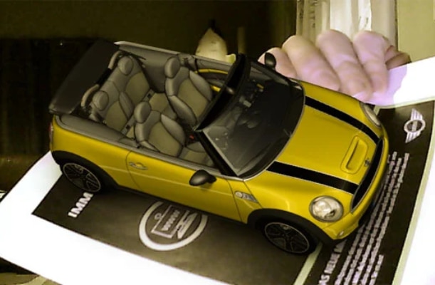 A yellow MINI automobile is displayed in 3D using augmented reality (AR) over a 2D magazine advertisement. Original image by technabob located here: https://technabob.com/blog/2008/12/17/mini-augmented-reality-ads-hit-newstands/